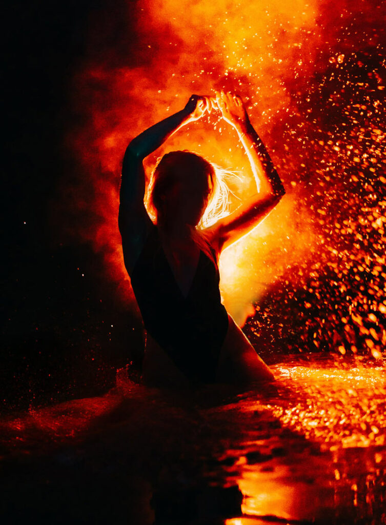Front-facing self-portrait standing in a forest lake at night, silhouetted by warm backlight and a bright reflection on the rippling water surface