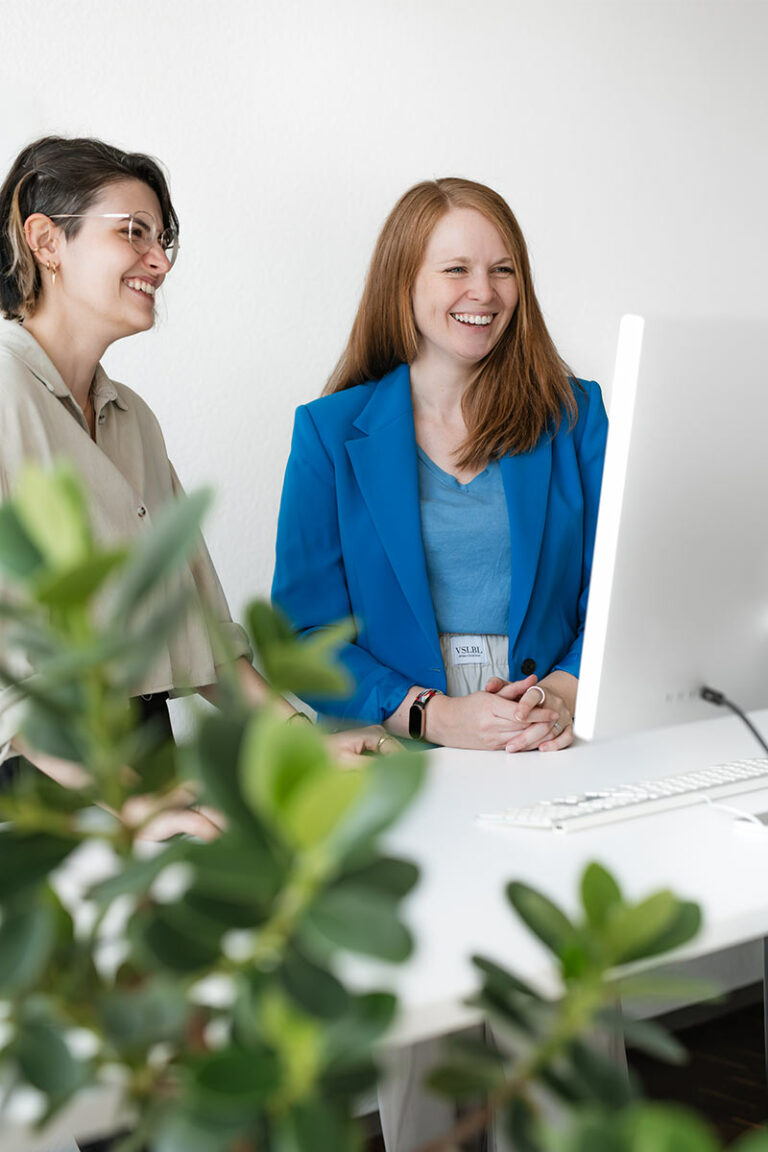 Businessfotos in der Agentur Lehnen Markenreich in Ulm, zwei Frauen stehen lachend an einem hochgefahrenen Schreibtisch und blicken auf den Bildschirm