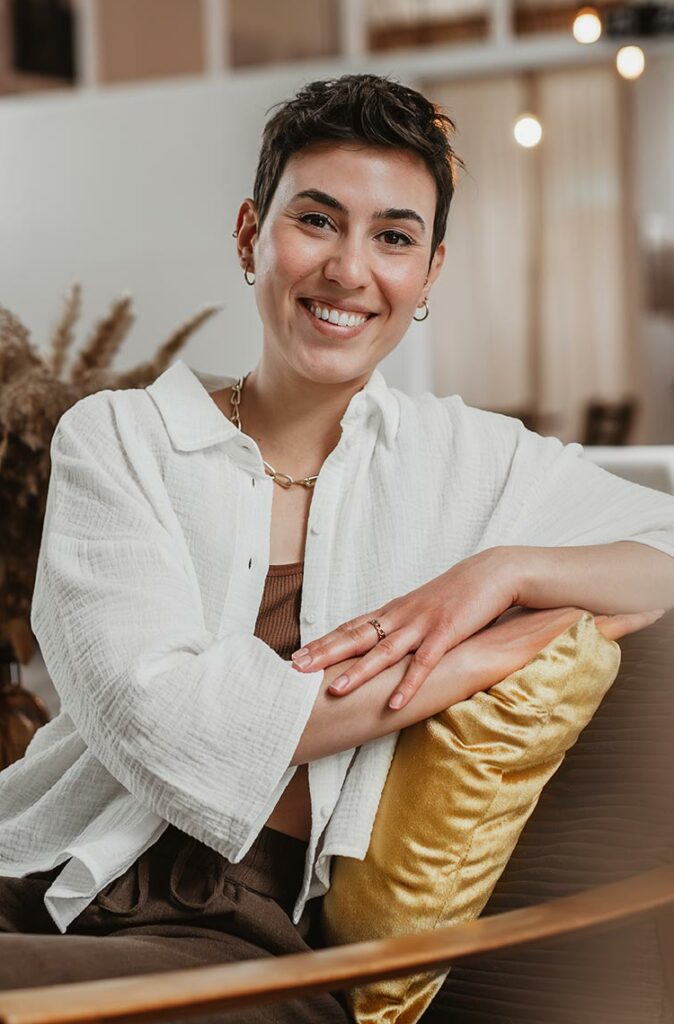 Kathrin von Deepshit, sitzend auf einem holz-Stuhl in Braun mit einem goldenen Kissen (ihre Branding Farben), sie trägt ein weißes leichtes Hemd und lächelt in die Kamera